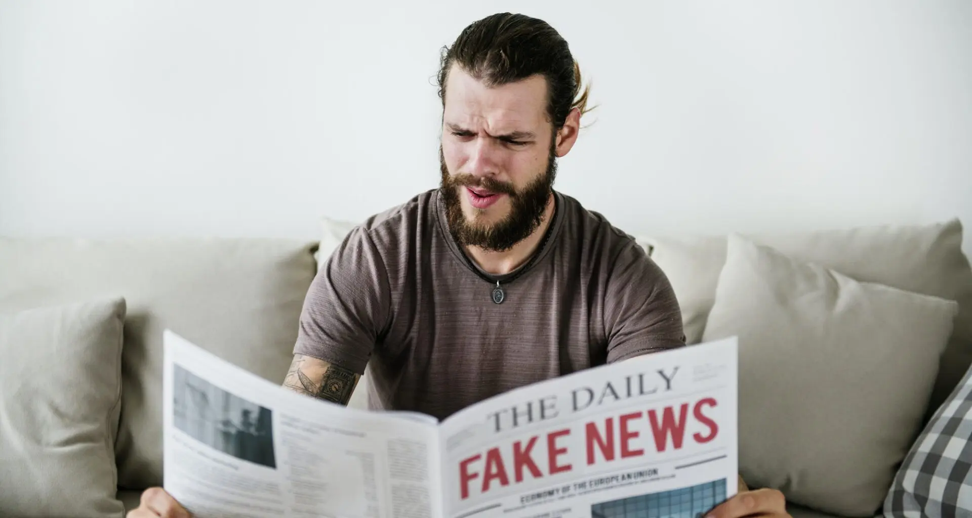 A man with a beard, sitting on a couch, appears perplexed or frustrated while reading a newspaper with the headline "FAKE NEWS."