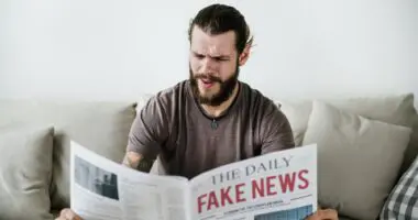 A man with a beard, sitting on a couch, appears perplexed or frustrated while reading a newspaper with the headline "FAKE NEWS."