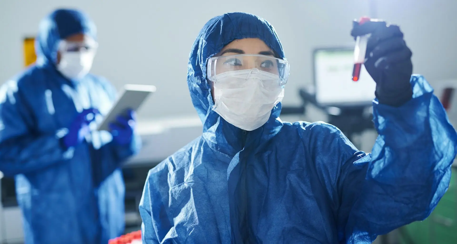 Two healthcare professionals in blue protective suits, masks, and gloves, working in a laboratory.