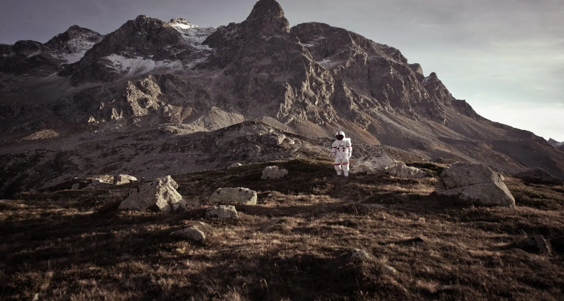A person in a white space suit stands on a rocky terrain with mountains in the background, evoking the feel of a space colony.