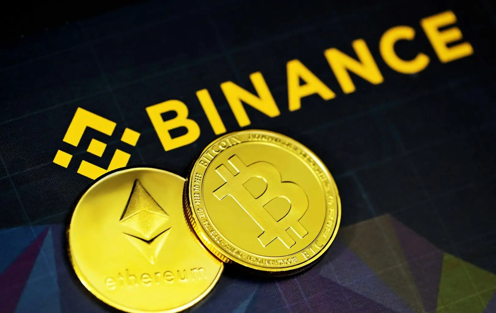 Two gold-colored cryptocurrency coins with 'Bitcoin' and 'Ethereum' logos on them, placed on a surface with the word 'BINANCE' in the background.
