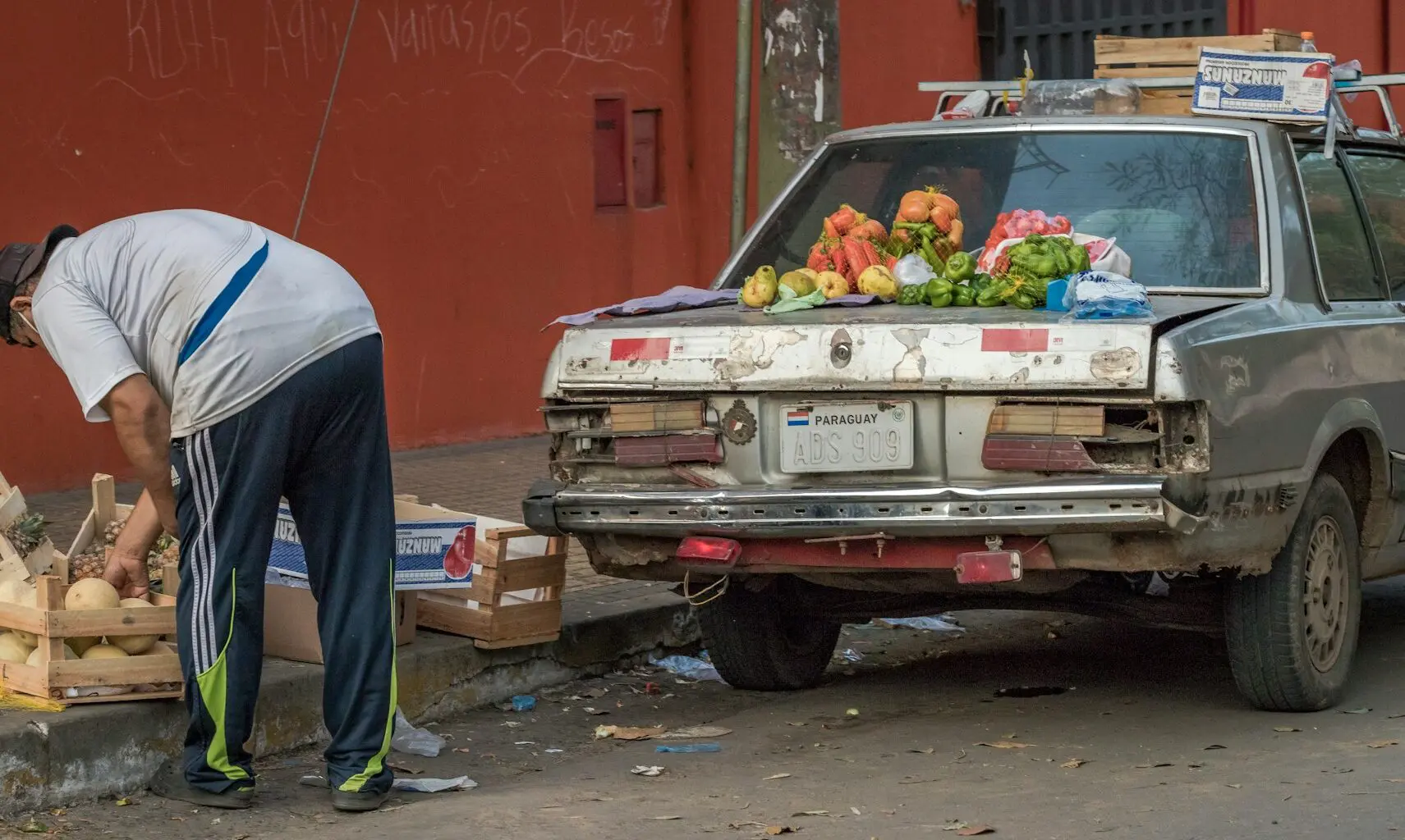 A person is bending over to sort items beside an old, damaged car with various fruits and vegetables on its trunk, parked on a street.