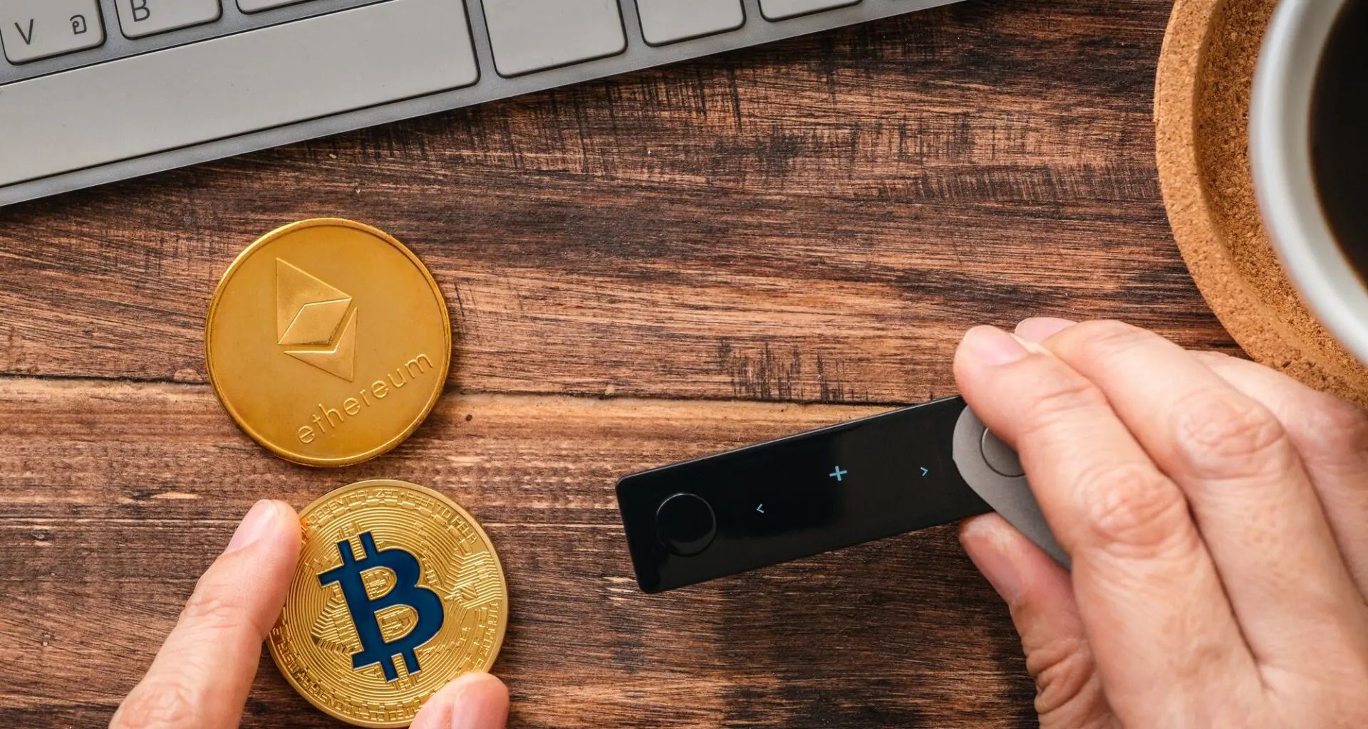 A person's hand holding a hardware wallet next to a Bitcoin and an Ethereum coin on a wooden desk, with a keyboard and a cup of coffee nearby.