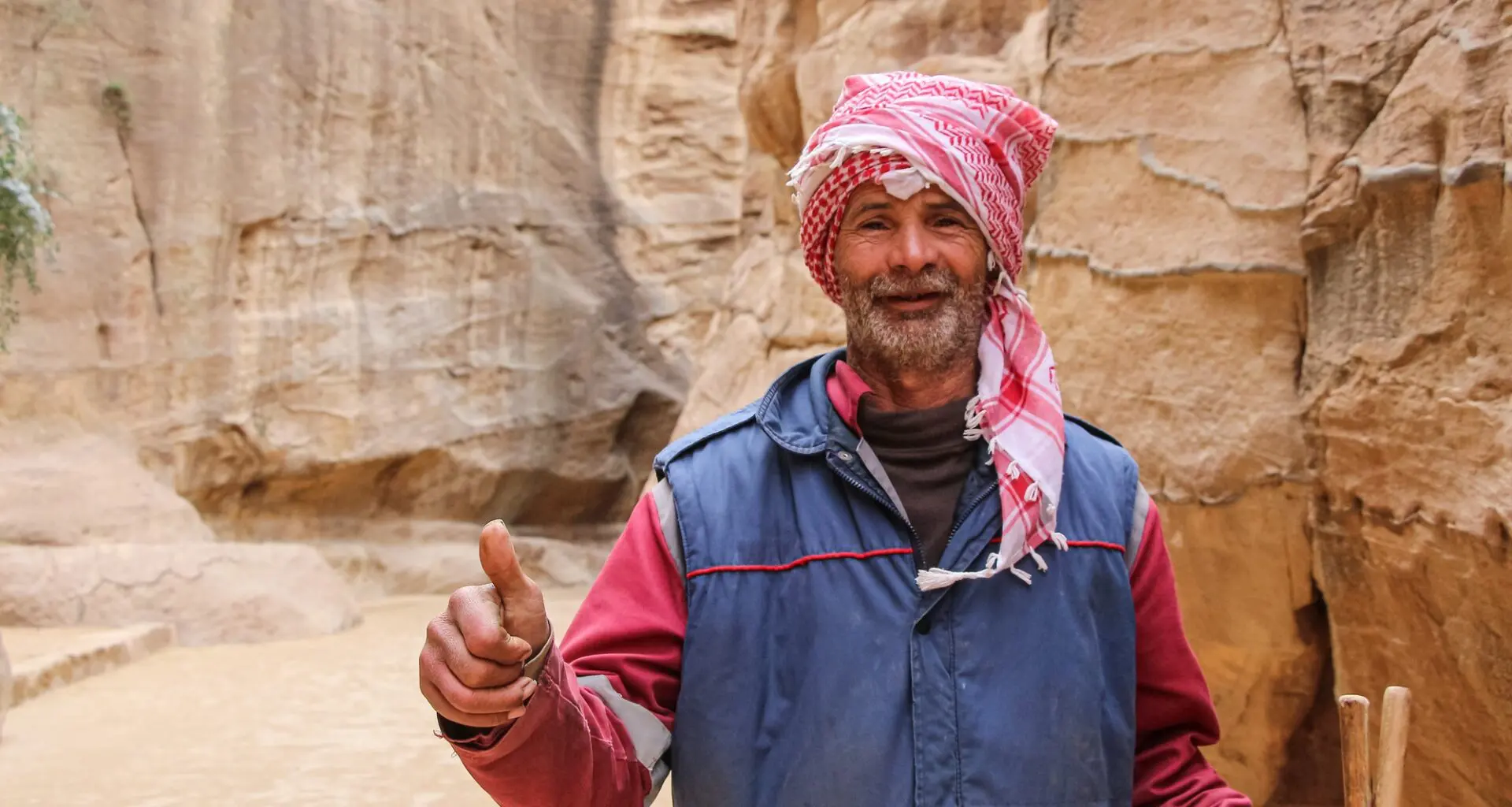 A man in a red and white headscarf, typical of Jordanian attire, standing before a rocky desert background.