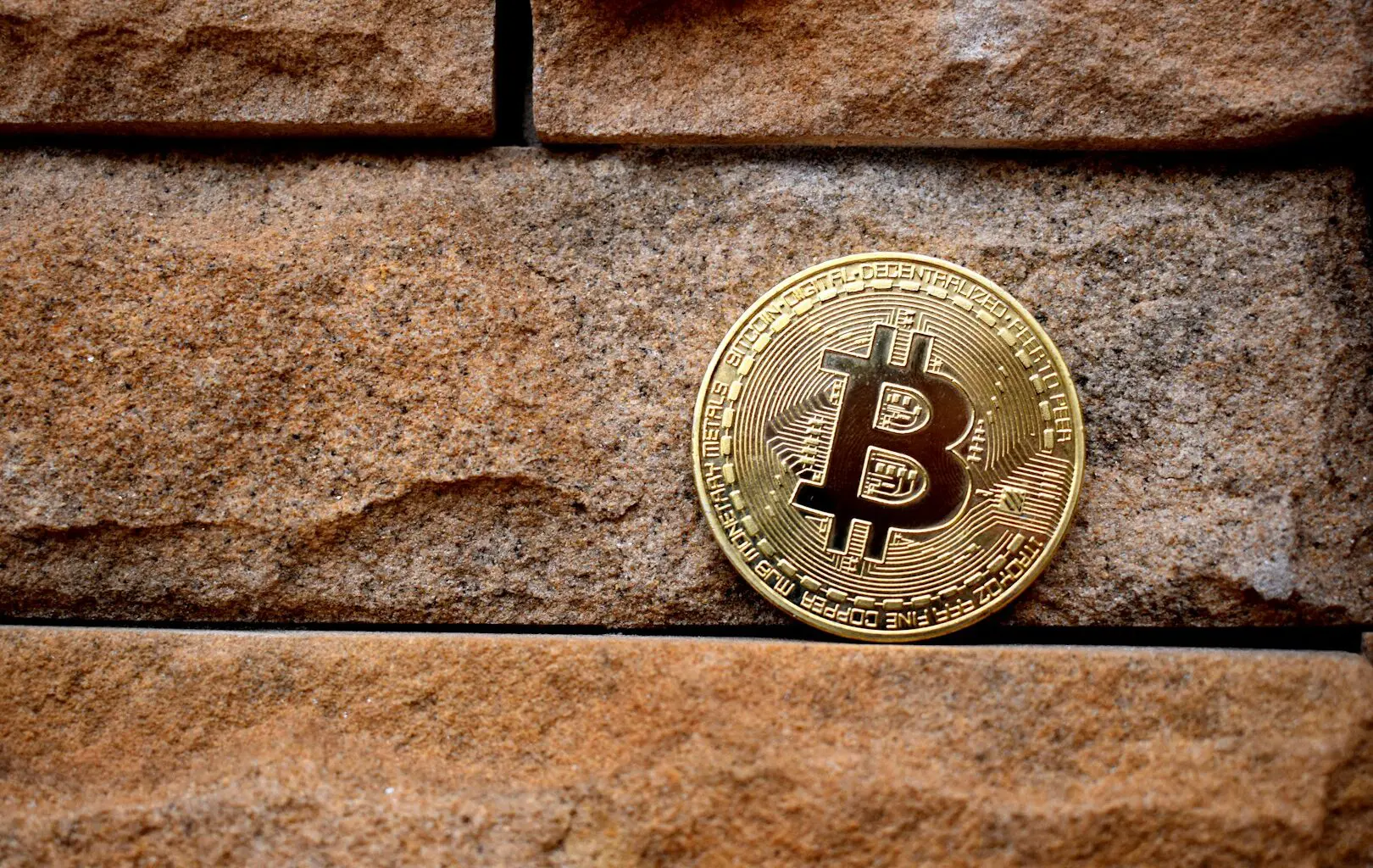A gold Bitcoin sits on top of a red brick wall.