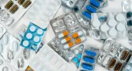 The image shows a pile of colorful pills and capsules in blister packs, with text referencing a pharmaceutical company and potential cost savings.