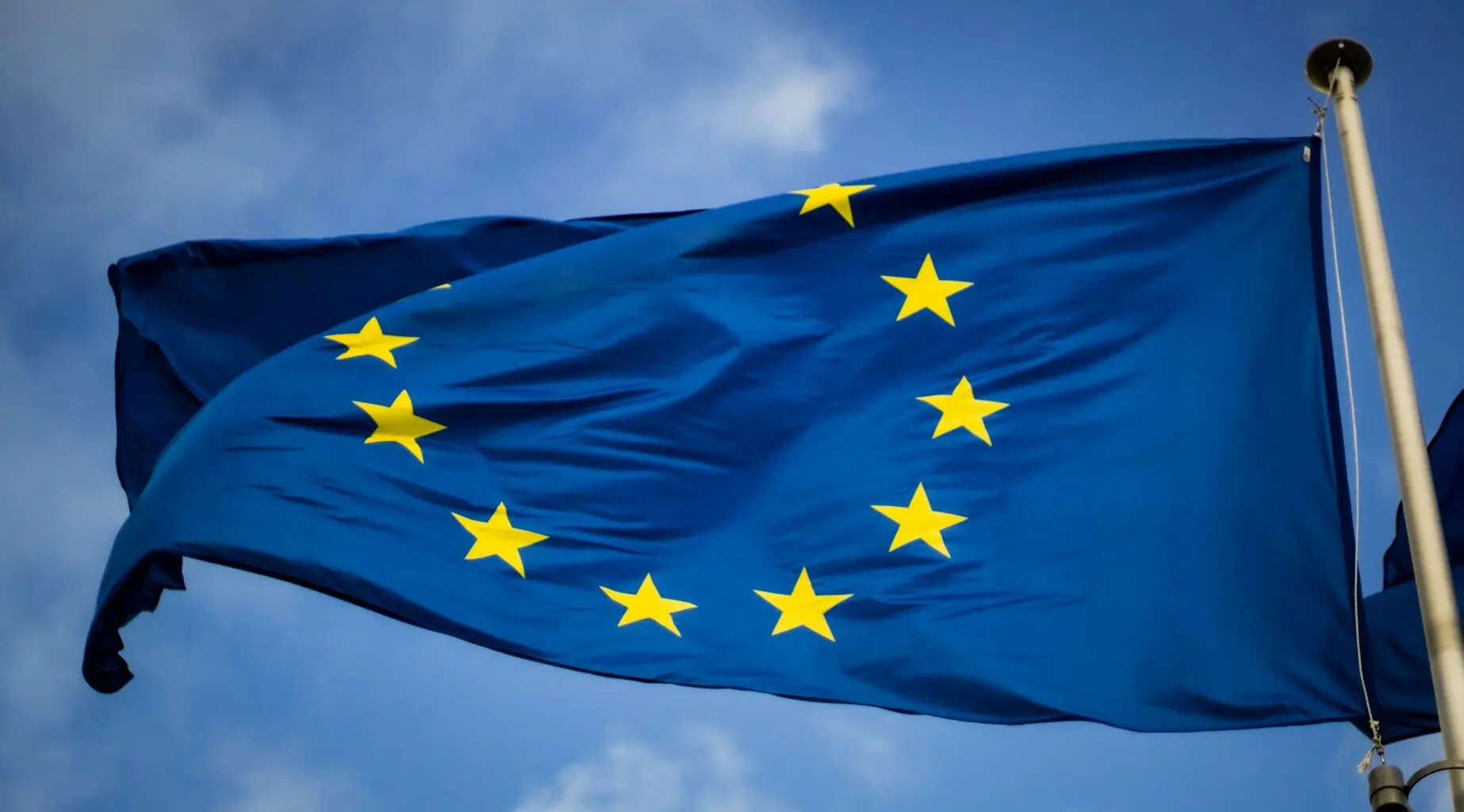The image depicts the flag of the European Union (EU), featuring a circle of twelve gold stars on a blue background, fluttering against a partly cloudy sky.