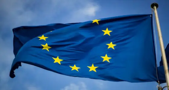 The image depicts the flag of the European Union (EU), featuring a circle of twelve gold stars on a blue background, fluttering against a partly cloudy sky.