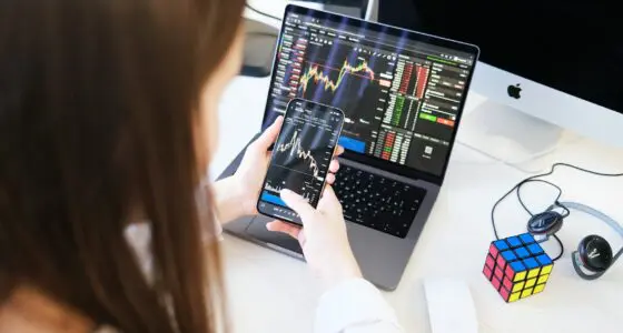 A person looking at a yield farming app on a smartphone with a laptop showing cryptocurrency charts and a Rubik's Cube on the desk.