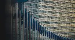 A row of European Union flags waves outside a modern glass-fronted building. The flags feature the distinctive blue background with yellow stars, symbolizing unity and cooperation among EU member countries.