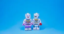 Two LEGO robots stand side by side against a blue background, holding hands. One robot has pink details while the other has red accents, symbolizing a relationship.
