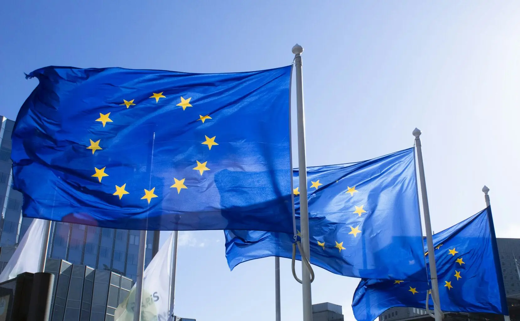 Three European Union flags flutter in the wind against a clear blue sky, with modern office buildings in the background. The flags are blue with a circle of twelve gold stars, symbolizing unity and harmony among the peoples of Europe.