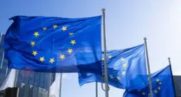 Three European Union flags flutter in the wind against a clear blue sky, with modern office buildings in the background. The flags are blue with a circle of twelve gold stars, symbolizing unity and harmony among the peoples of Europe.