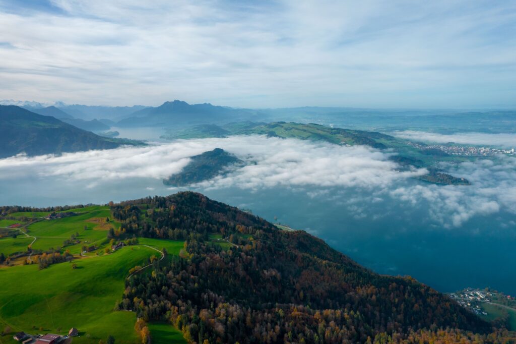 An aerial view from Zugerberg showcasing the expansive Lake of Zug partially veiled by clouds, with the city of Zug, Switzerland, visible in the distance, surrounded by lush greenery and the silhouette of mountains on the horizon.