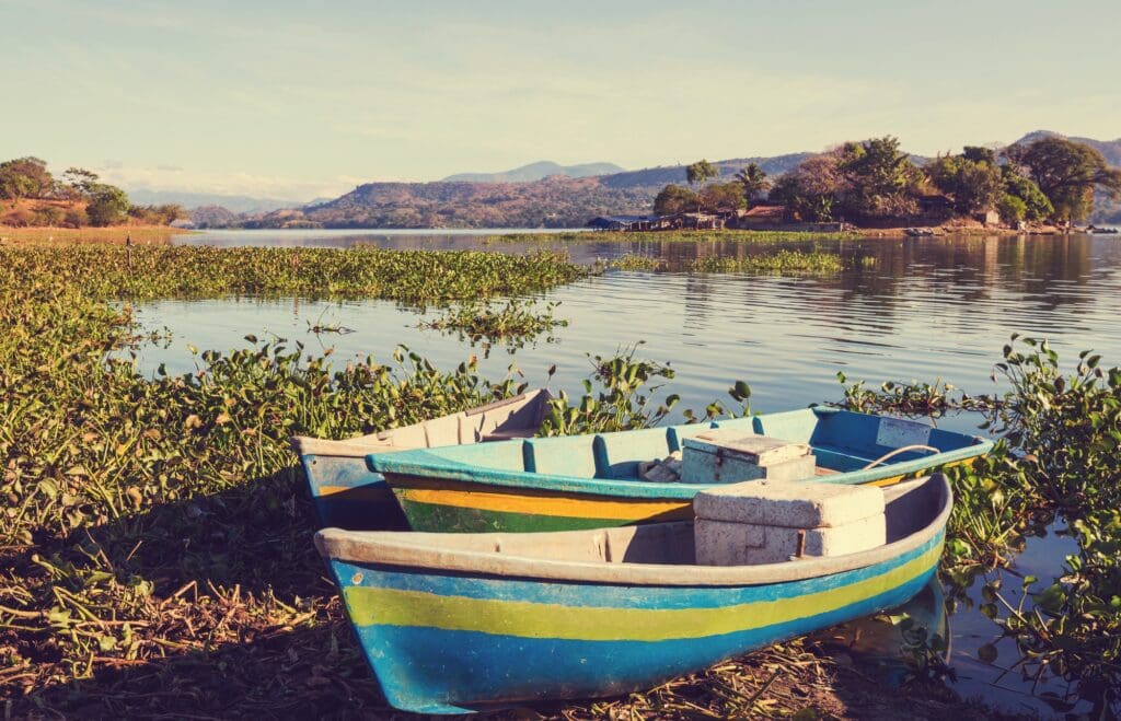 A colorful wooden boat moored at the lush shoreline of a lake in El Salvador, with tranquil waters and a backdrop of distant mountains under a clear sky.