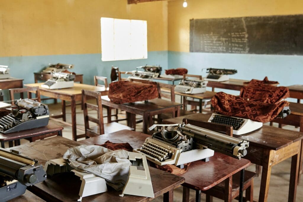 A classroom with vintage typewriters on wooden desks and a chalkboard in the background.




