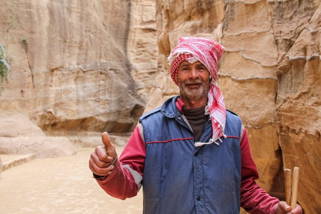 A man in a red and white headscarf, typical of Jordanian attire, standing before a rocky desert background.